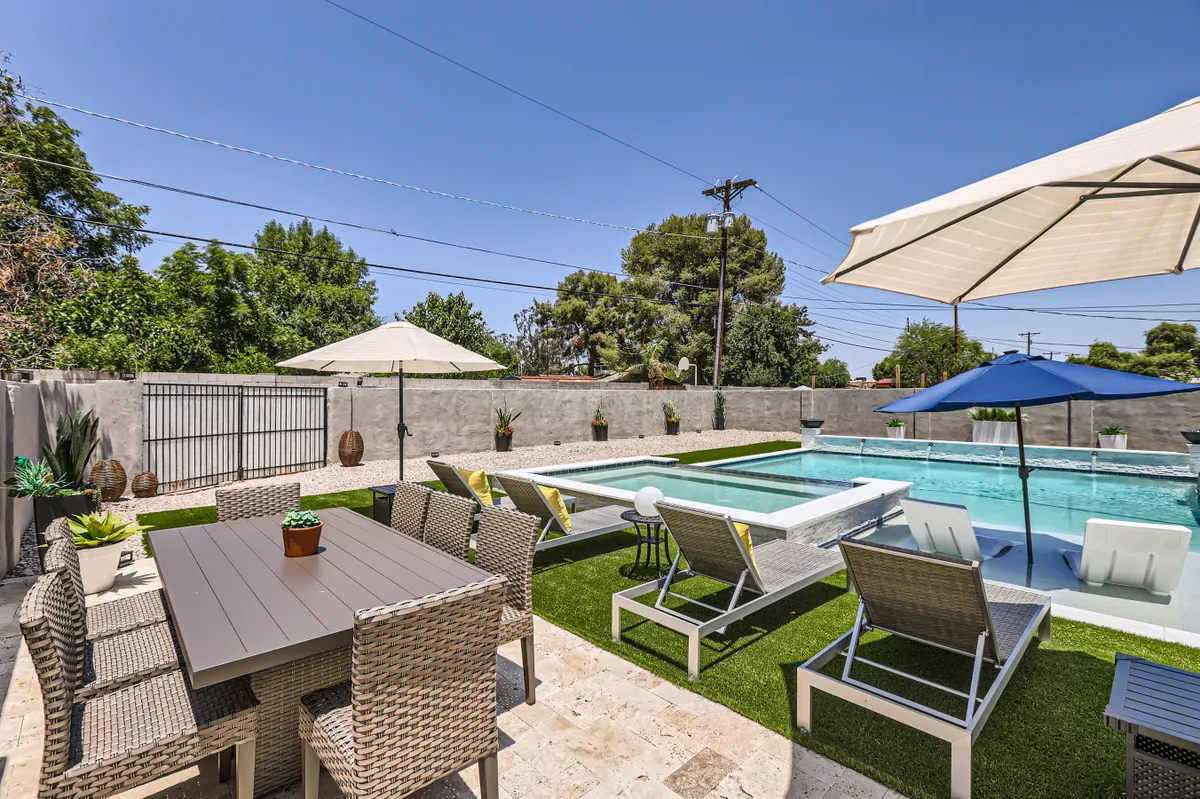 This exterior shot captures the backyard’s pool in the foreground, flanked by an outdoor dining table, lounge chairs, and umbrellas. The scene conveys a cheerful and social vibe, perfect for outdoor dining, celebrations, or casual lounging beside the water.