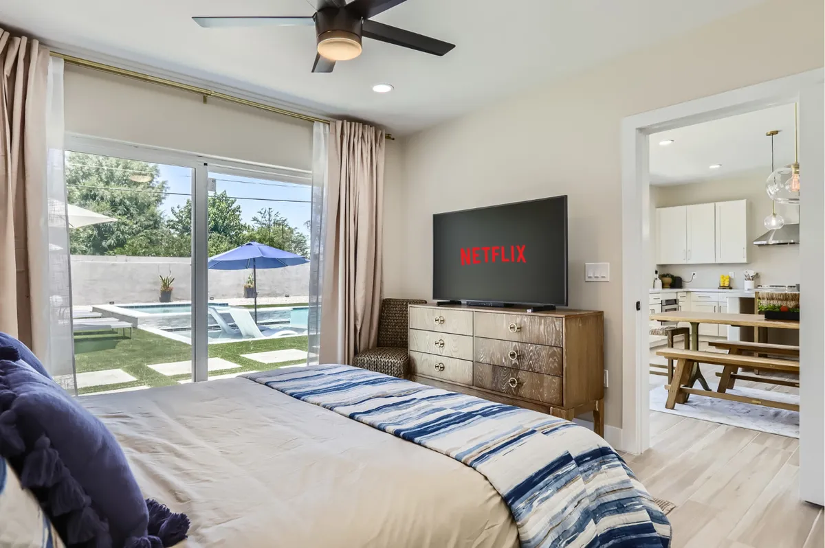 This interior image features a guest room with a striped blue and white bed, a large TV displaying Netflix, and a window that offers a glimpse of the pool and patio area outside. The wooden dresser and relaxed setup highlight a comfortable, functional space well-suited for both rest and remote work.