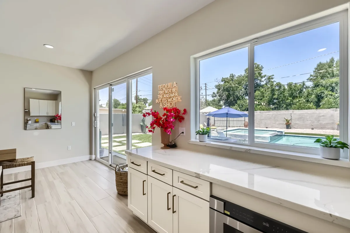 A kitchen-centric entertaining space with a large island countertop, bar stools, and large windows that flood the area with natural light. A decorative sign and floral arrangement add playful, stylish touches to the vibrant setting.