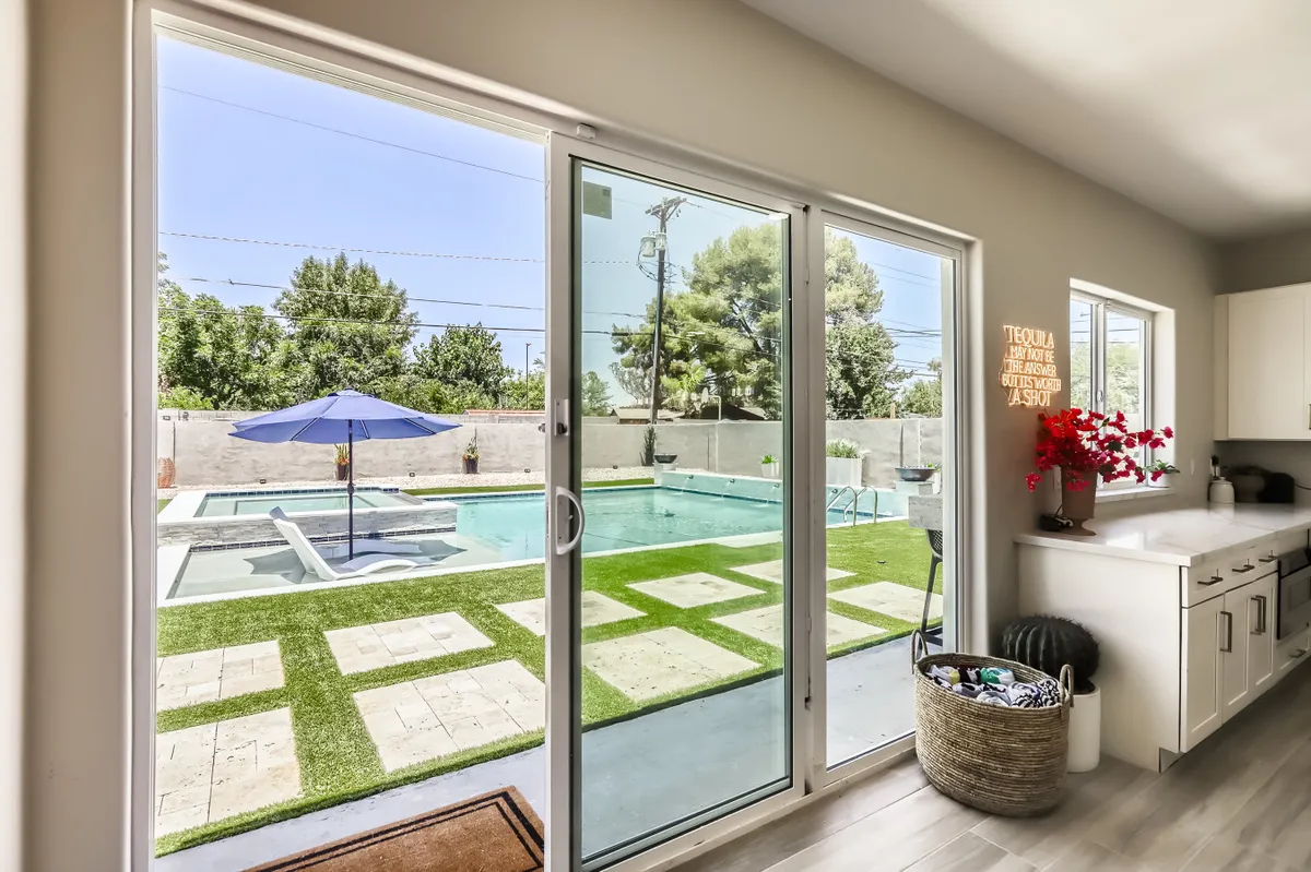 An inviting view that looks out from an interior living area through sliding glass doors towards the pool, umbrella, and outdoor seating. The room is filled with natural light and features a decorative plant, enhancing the indoor-outdoor connection.