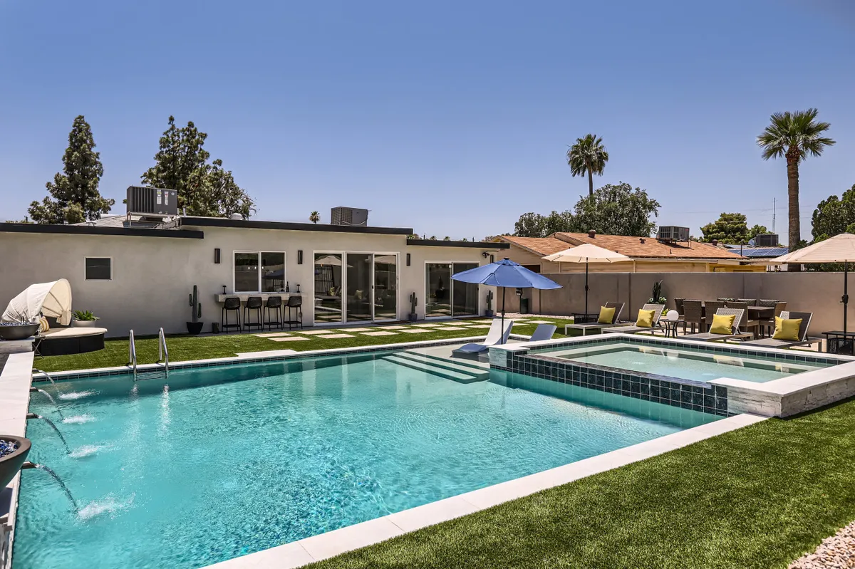The image provides a wider view of the backyard, emphasizing both the swimming pool and the extra-large jacuzzi (spa area). Umbrella-shaded seating and additional lounge chairs are visible, offering cues for relaxation and gathering spaces, while a subtle sunset glow evokes warmth and evening enjoyment.
