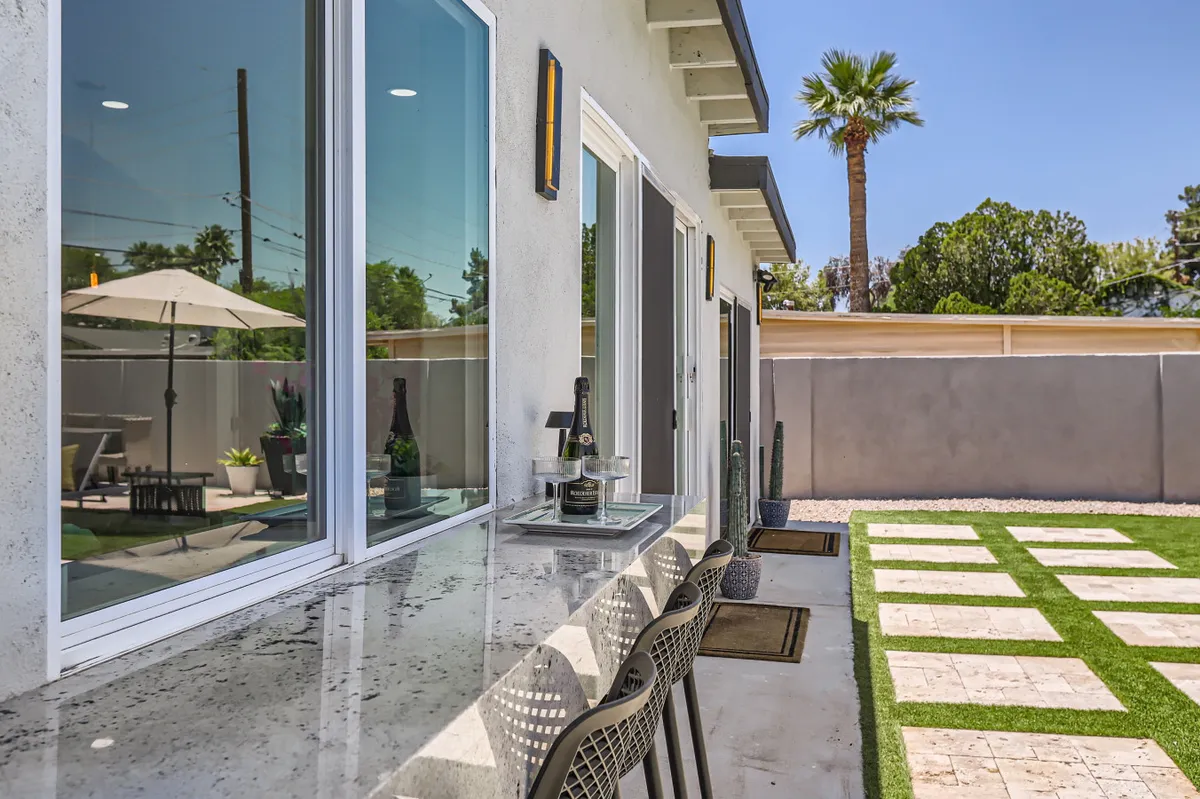 An inviting outdoor bar area with champagne bottles, bar seating, grassy lawn, a palm tree, and a patio umbrella. The scene is bright and festive, capturing the essence of entertaining or enjoying a morning coffee in a lush, resort-like environment.