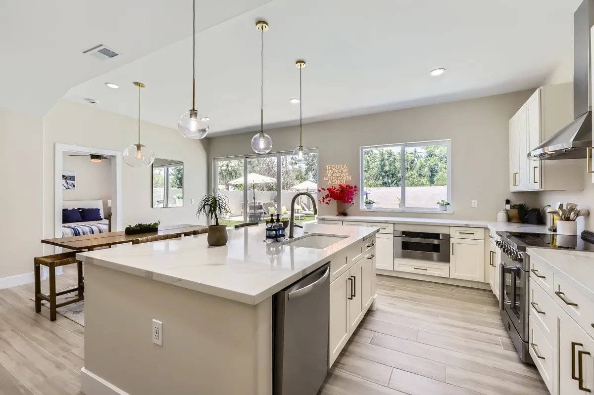 A bright, modern kitchen filled with natural light, featuring a large kitchen island, sleek modern appliances, and a decorative plant with vibrant red flowers. The space feels open and inviting, suggesting a lively environment perfect for gathering before heading out or enjoying a sustainable meal at home.